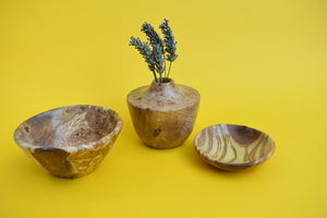 A wooden bowl, a small wooden dish and a wooden vase with dried lavender inside on a yellow background. 