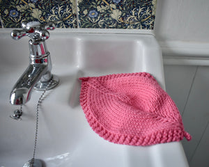 A pink leaf shaped hand knitted cotton cloth on the edge of a white sink. Next to the cloth is a tap and there are flowered tiles in the background. 