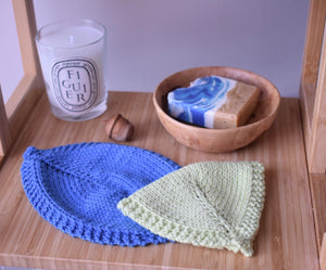 A green leaf shaped hand knitted cloth lies on top of a blue leaf shaped cloth on a wooden shelf. In the background there is a candle and wooden acorn and a wooden bowl with some coloured soap in it. 