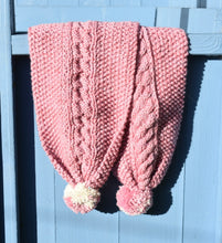Load image into Gallery viewer, A pink hand knitted scarf is draped over a light blue bench. The scarf has cute pom poms. 
