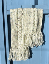 Load image into Gallery viewer, A cream hand knitted scarf speckled with colour and tassels hangs over a light blue bench. 
