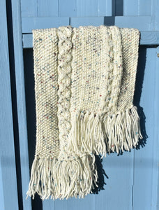 A cream hand knitted scarf speckled with colour and tassels hangs over a light blue bench. 