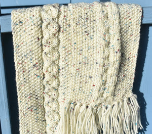 A close up of a cream scarf speckled with colour and with a cable pattern running down the centre. 