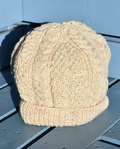 AN oatmeal coloured hand knitted hat with a cable pattern sitting on a light blue bench. 