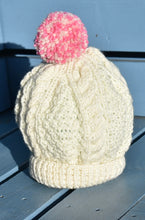 Load image into Gallery viewer, A cream hand knitted child hat with a pink and cream pom pom sitting on a blue bench. 
