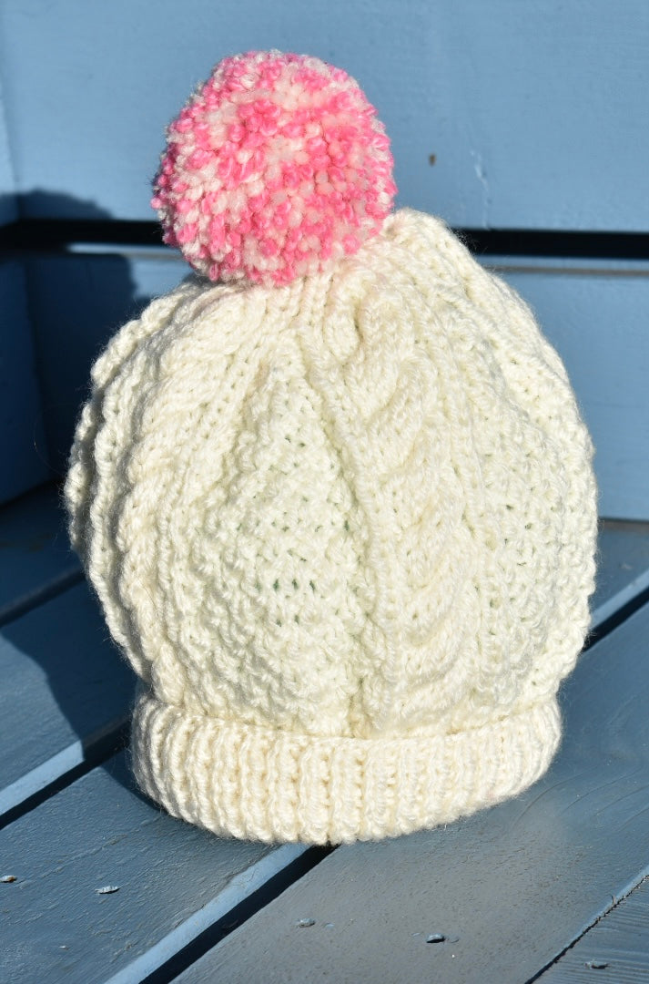 A cream hand knitted child hat with a pink and cream pom pom sitting on a blue bench. 