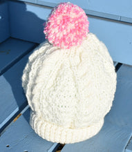Load image into Gallery viewer, A close up of a childs cream hand knitted hat with a pink and cream pom pom. 
