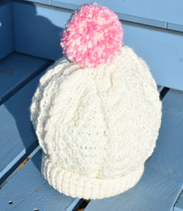 A close up of a childs cream hand knitted hat with a pink and cream pom pom. 