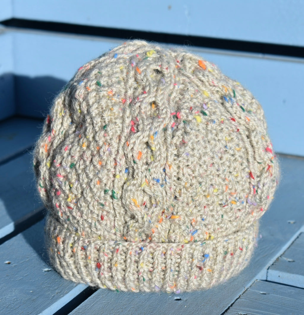 An oatmeal coloured hand knitted hat with a colourful speckled effect all over. The hat has as cable pattern and is sitting on a light blue bench. 