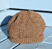 Load image into Gallery viewer, A dark brown hand knitted hat with a cable pattern sitting on a light blue bench. 
