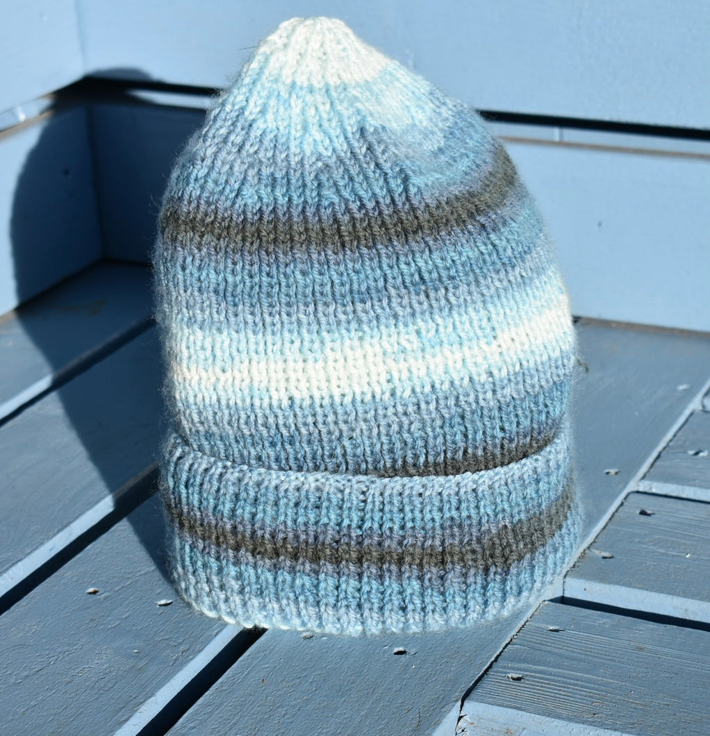 A hand knitted hat sitting on a light blue bench. The wool has created a stripe patter made up of different blues and dark grey. 