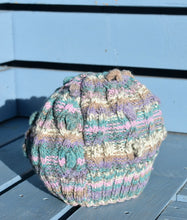 Load image into Gallery viewer, A multi coloured hand knitted hat sits on a light blue bench. 
