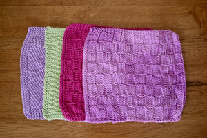 Four brightly coloured square cotton hand knitted cloths lying on a wooden background each slightly overlapping. 