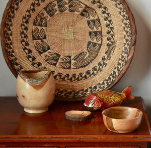 A set of three wooden bowls of different sizes on a wooden table with a metal carp childrens toy and a large round African basket in the background 