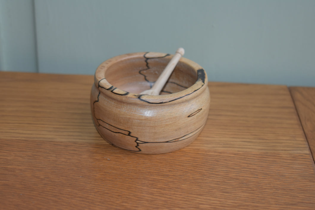 A small handturned wooden bowl with a small wooden scoop in it sitting on a wooden bench with a pale green background.