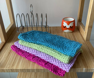 A set of four brightly coloured hand knitted cotton cloths , folded in half and piled up on a wooden shelf with a silver toast rack and fox egg cup in the background. 